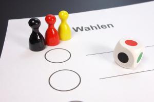 Zu sehen ist ein Wahlzettel mit mehreren Auswahloptionen zum Ankreuzen, ein Würfel mit den Farben Schwarz, Rot, Grün und drei Spielfiguren vom Rausschmeißer in den Farben Schwarz, Rot, Gelb